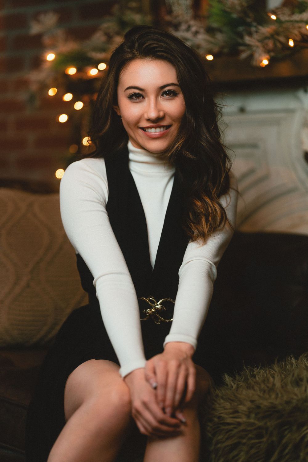 Smiling young woman in white turtleneck and black jumper seated near fireplace with warm string lights in Cincinnati