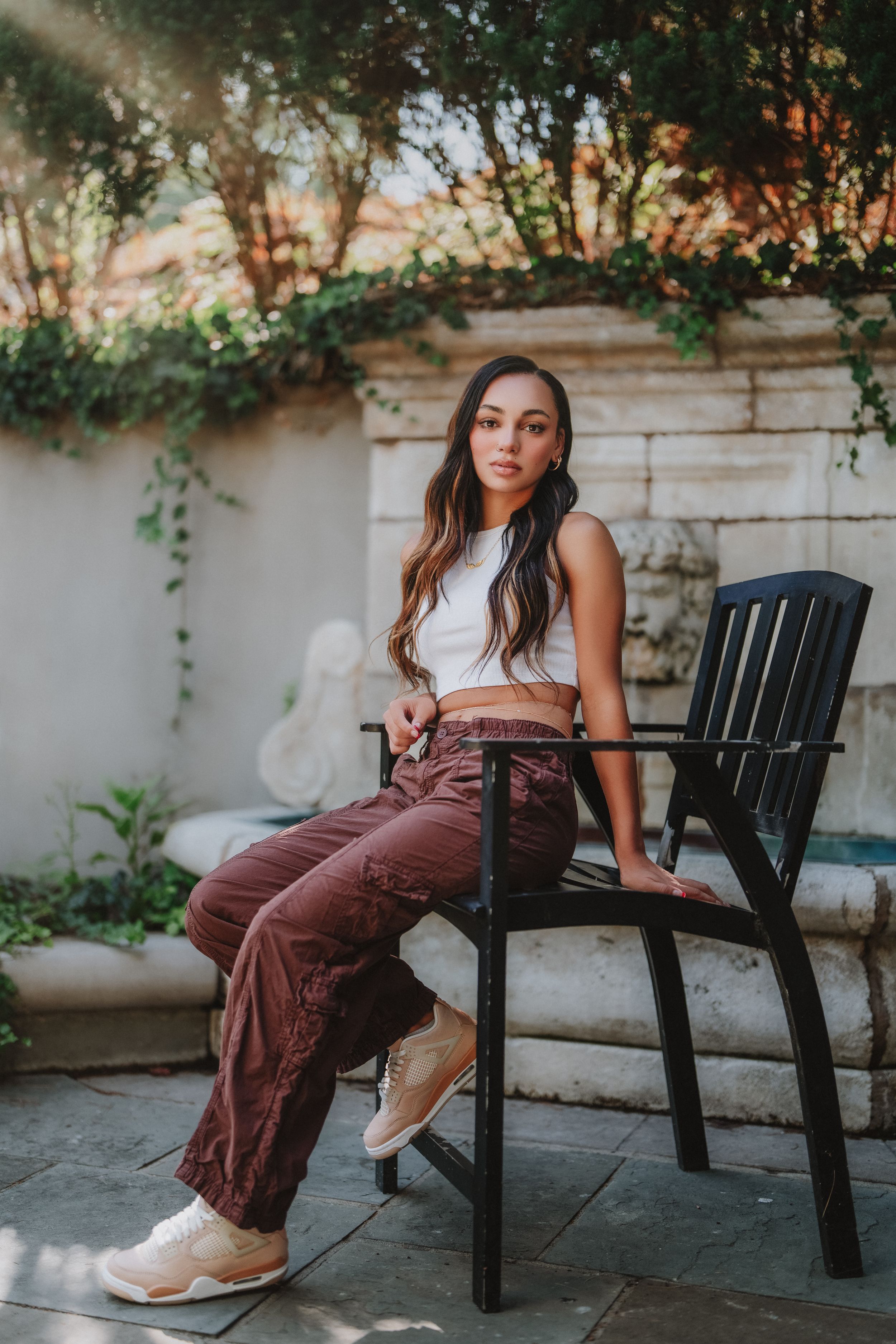 Young woman in white crop top and brown cargo pants sitting on black chair in garden patio setting in Cincinnati