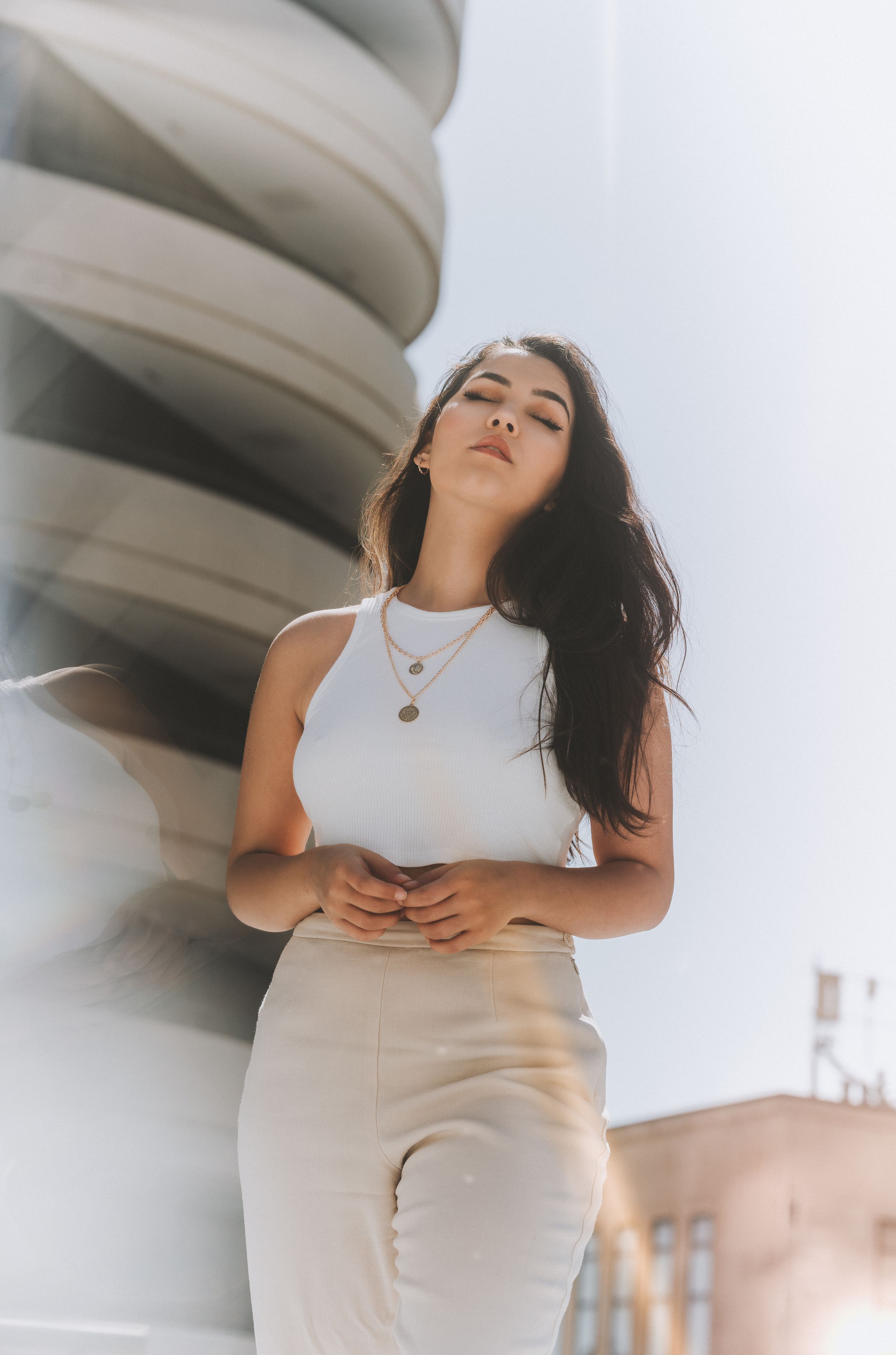 Young woman with long dark hair in white tank top posing near modern architectural structure outdoors for her senior session in Cincinnati