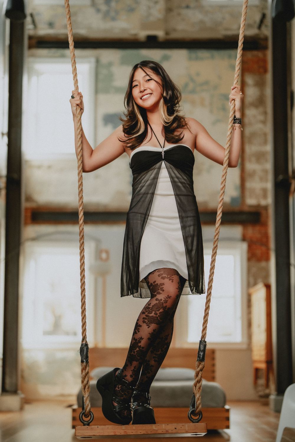 Smiling woman in black and white dress standing on wooden swing with rope in rustic loft space for her senior session in Cincinnati