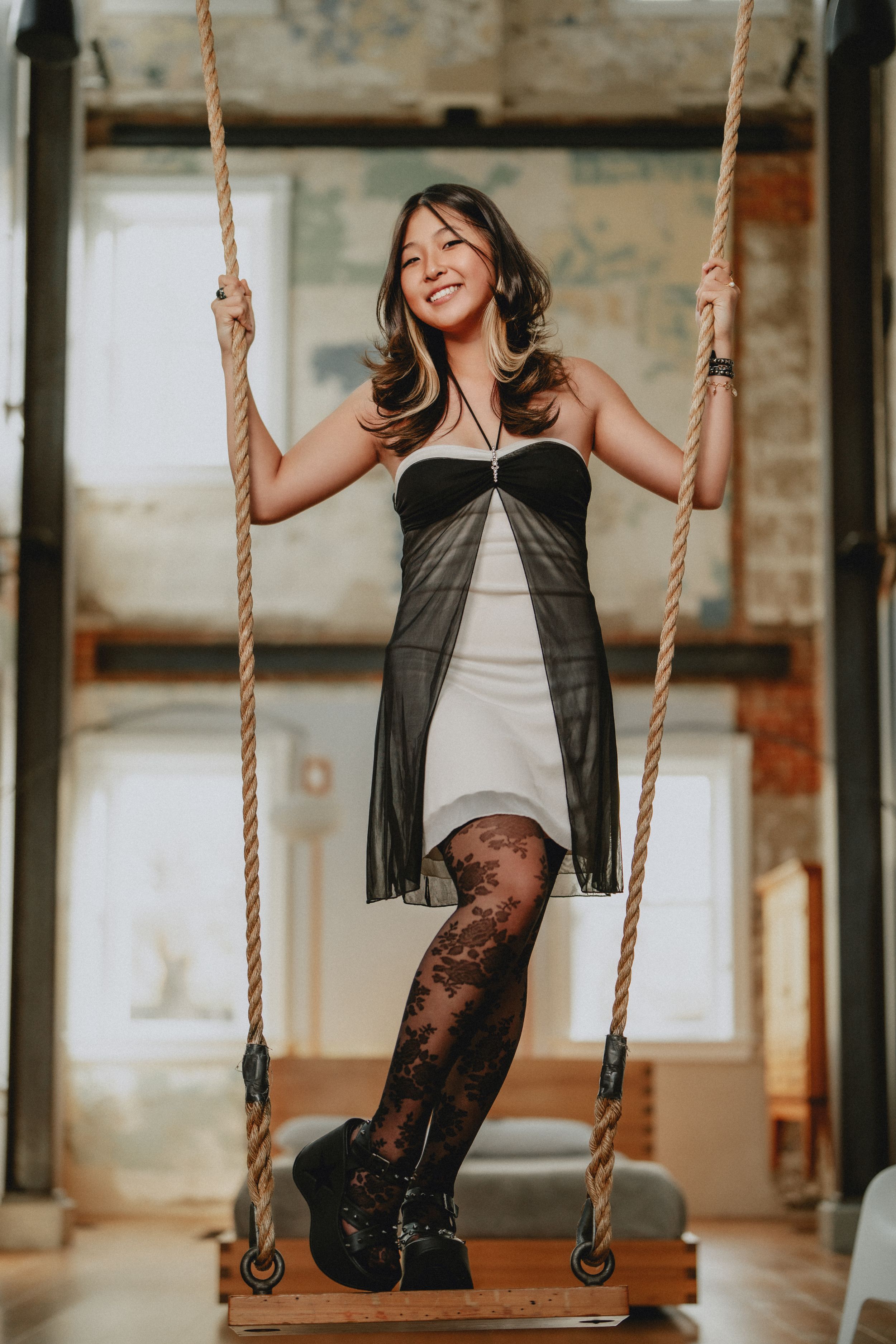 Smiling woman in black and white dress standing on wooden swing with rope in rustic loft space for her senior session in Cincinnati