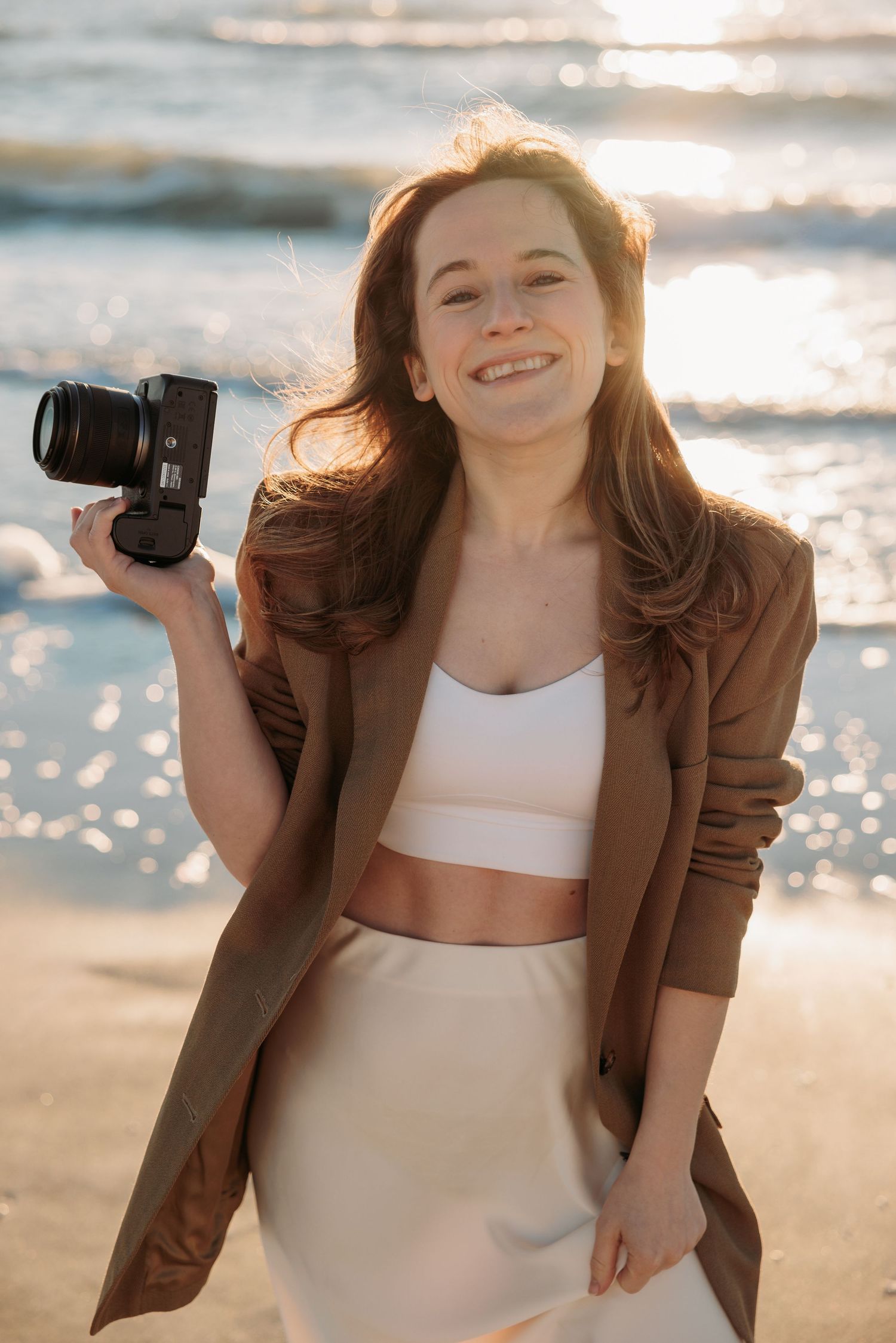 A woman in a plaid blazer holds up a vintage camera while smiling outdoors.