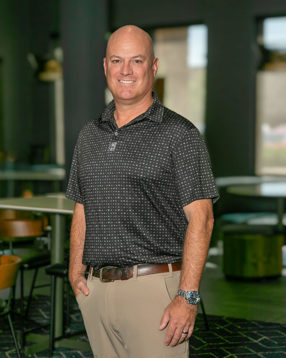 Professional portrait of someone in a dark polka dot shirt and khaki pants standing against an office background.