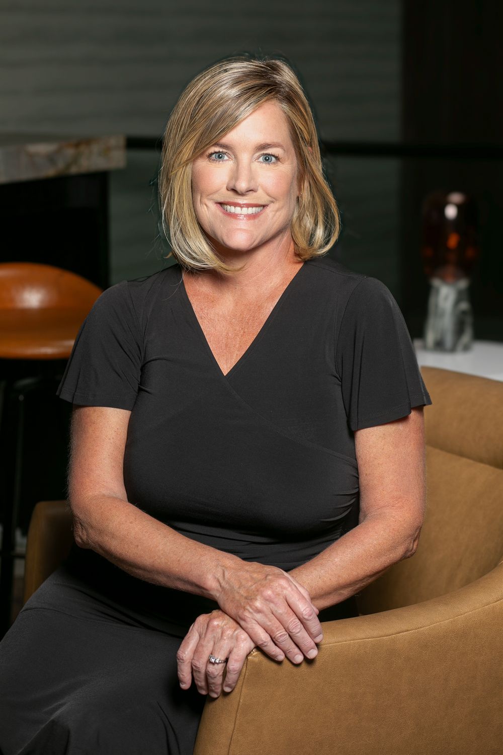 Professional business headshot of woman in black v-neck shirt seated in tan leather chair against dark background.