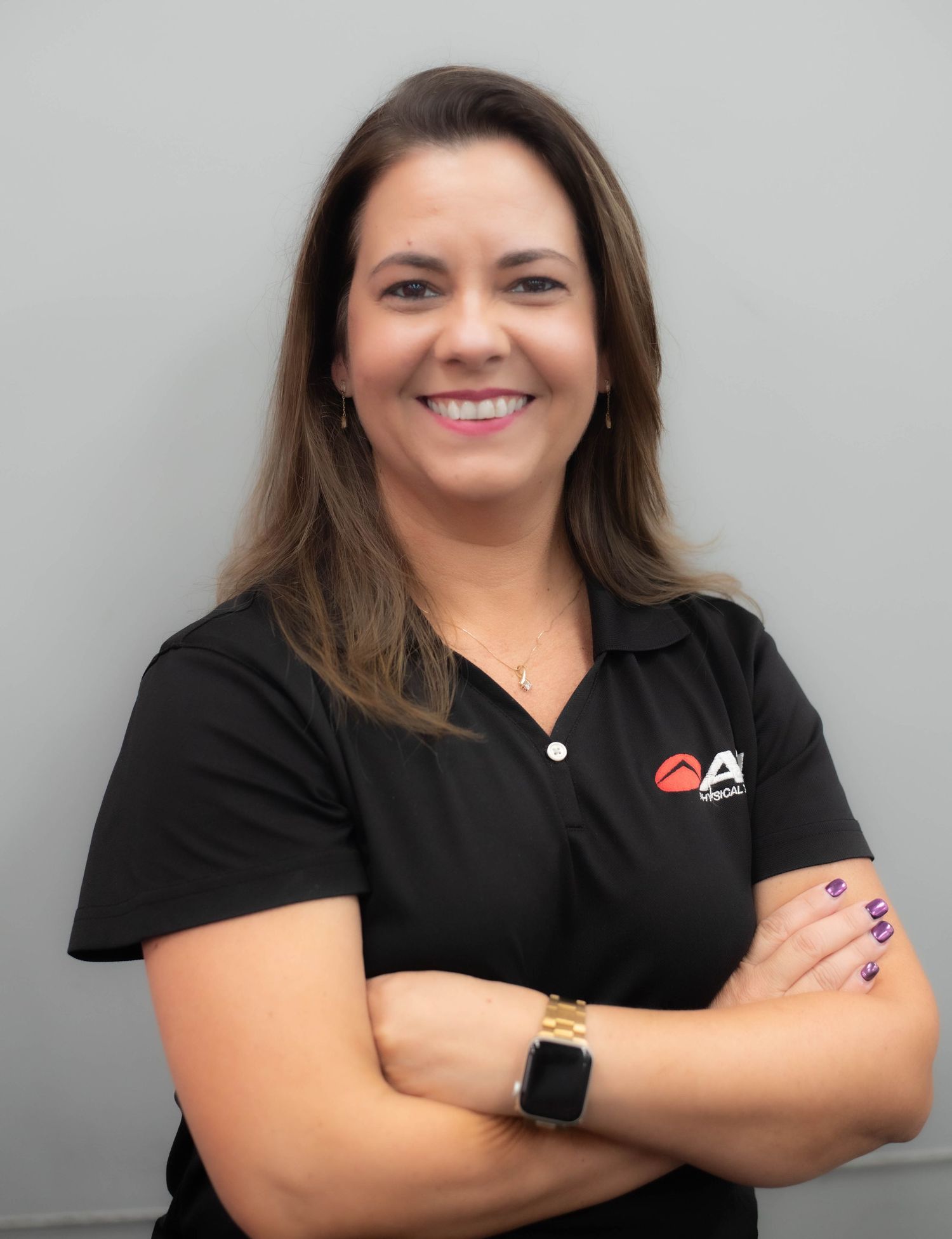 Woman in black polo shirt with logo smiles confidently with arms crossed against gray background.