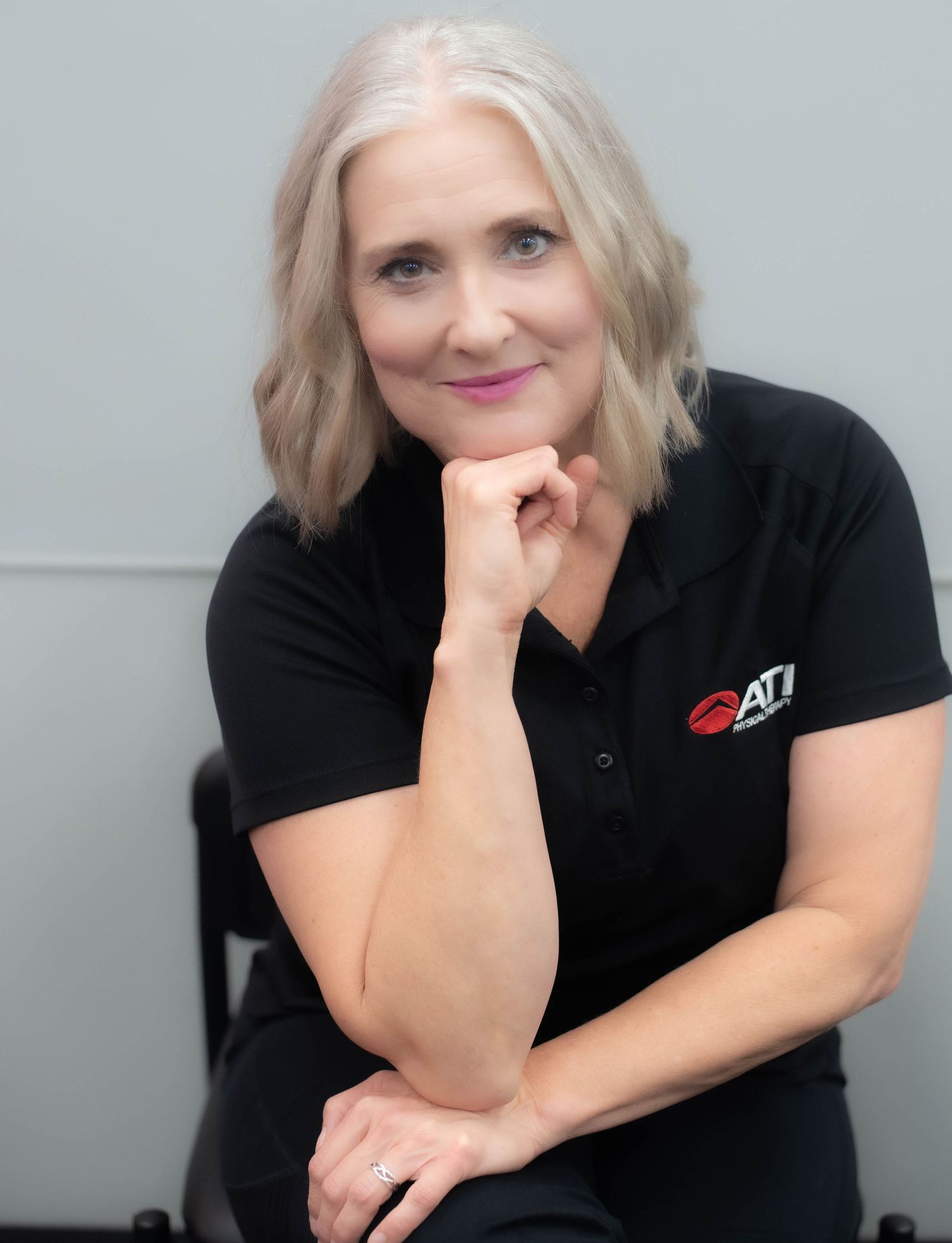 Woman with silver bob hair in black ATI polo shirt poses confidently with hand on chin, smiling warmly.