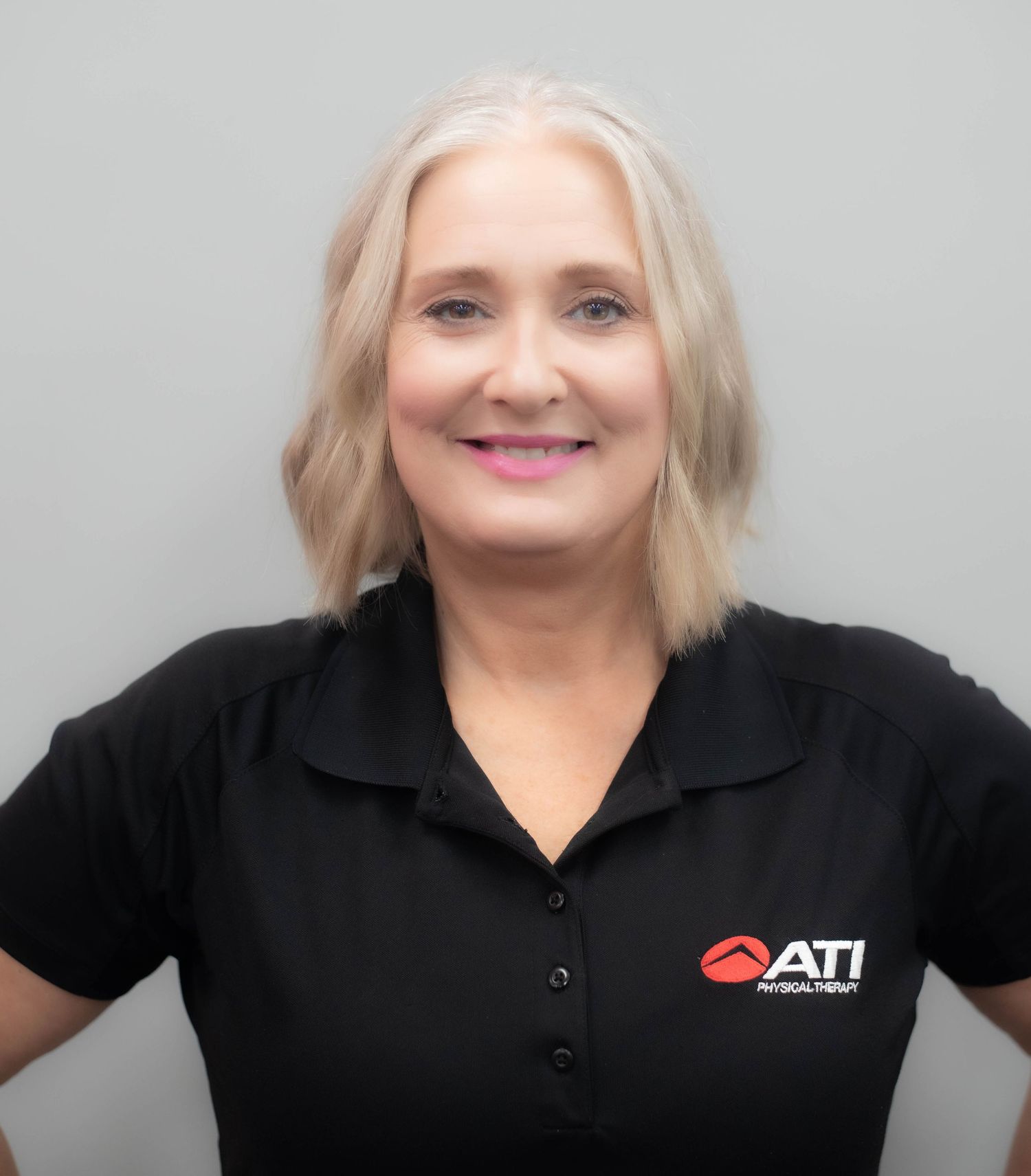 Smiling blonde woman in black ATI Physical Therapy polo shirt poses against gray background.
