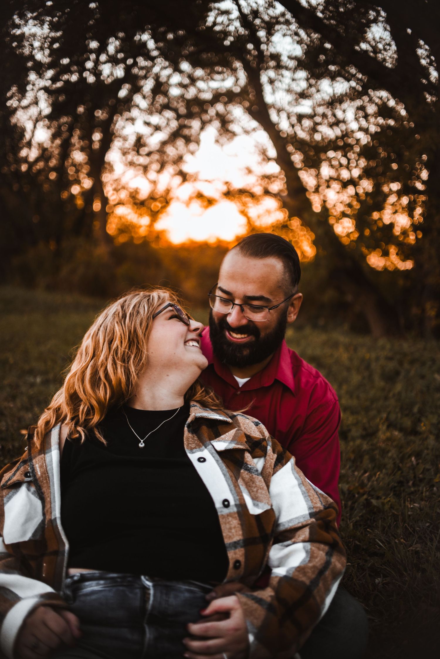 Romantic couple embraces and laughs together in sunset field with autumn trees silhouetted in background.