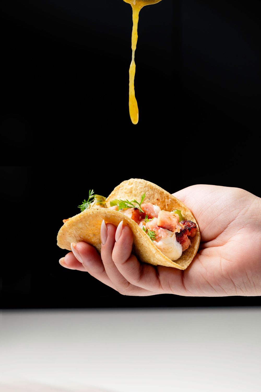 Yellow sauce dripping onto a mini taco held in hand against black background.