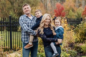 Family of four smiles together in front of autumn foliage and black iron fence.