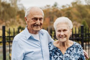 Senior couple embraces while smiling outdoors with fall colors in background.