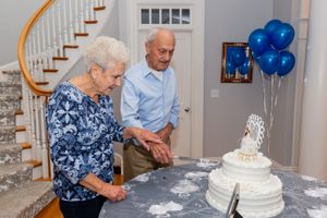 Senior couple looks at white tiered celebration cake with blue balloons in background.