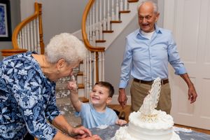 Three generations share a moment around an elegant white celebration cake near curved staircase.