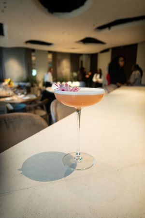 Elegant cocktail in a coupe glass with pink garnish sits on a white surface in a dimly lit setting.