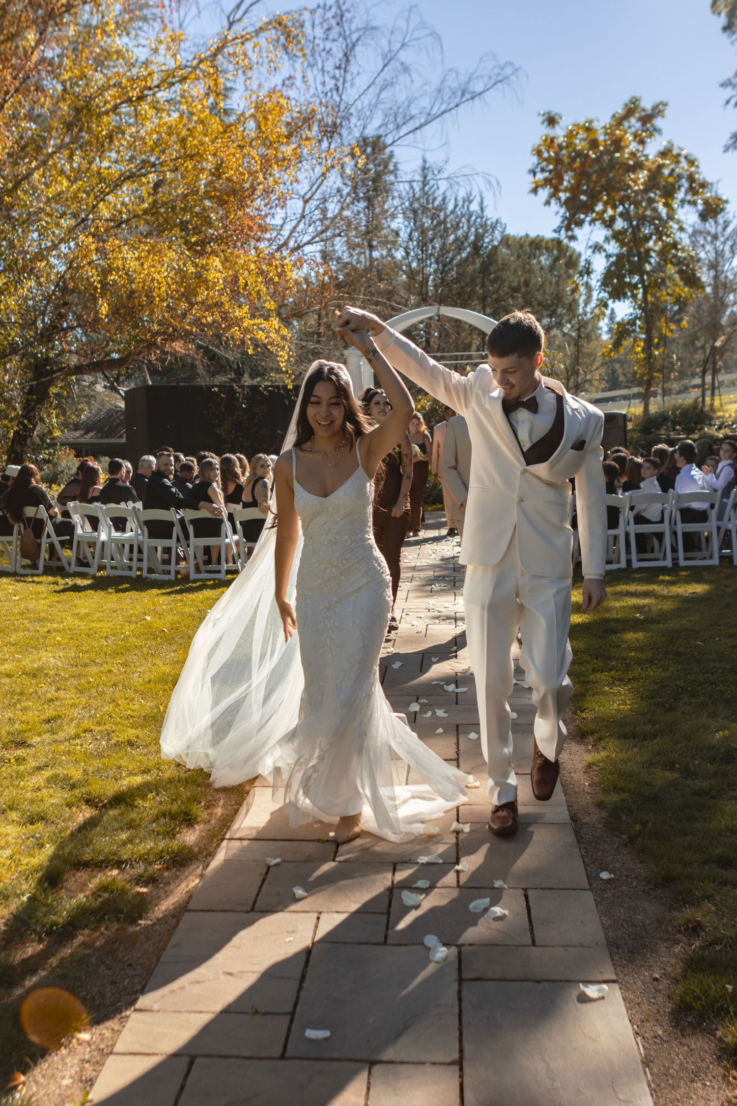 A married couple walks down the aisle on their wedding day. They're backlit, and he spins her as they walk. They're both in white.