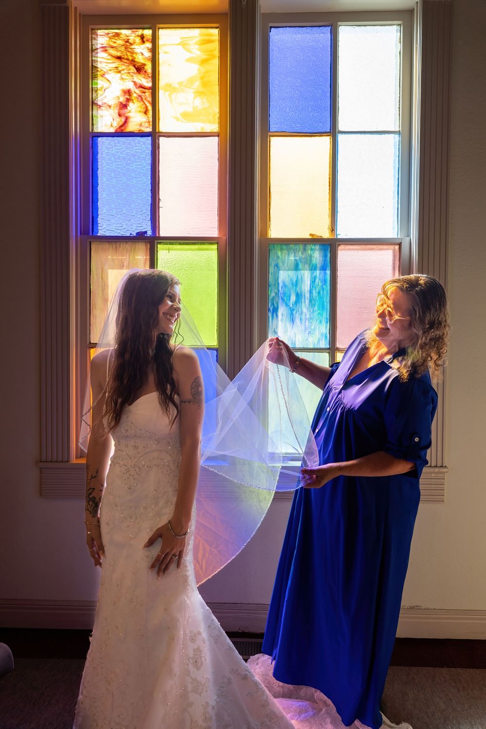 Bridesmaid in blue dress helps bride with veil before vibrant stained glass windows in chapel.