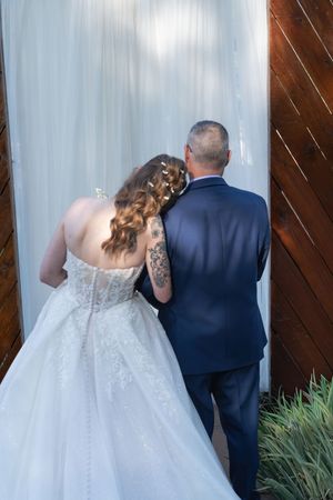 Bride in lace wedding gown and tattooed arm leans on groom in navy suit, viewed from behind near wooden wall.