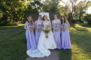 Bride in white gown with four bridesmaids in lavender dresses holding bouquets outdoors on green lawn.