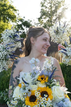 Smiling bride holding sunflower and white bouquet surrounded by floral arch in sunlit garden.