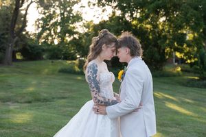 Bride with tattoos in white gown and groom in white suit touching foreheads in lush green garden during golden hour.