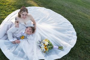Bride in white gown sits on grass with groom lying in her lap, bouquet of yellow sunflowers nearby.