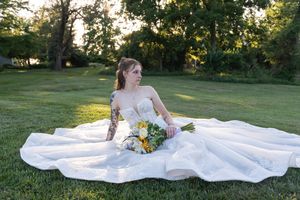 Bride in white gown with floral bouquet sitting on green grass outdoors, showcasing tattoos and elegant wedding dress.