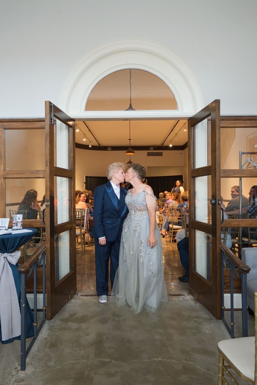 Two brides share a kiss in elegant attire at wedding venue doorway, one in navy suit, one in silver gown.