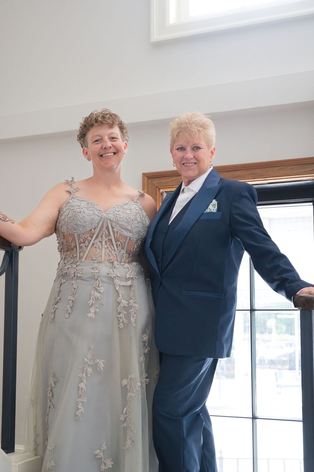 Two women in formal attire, one in a silver gown and one in a navy suit, posing on a staircase.