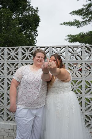 Two brides in white attire showing off wedding rings, smiling in front of decorative lattice wall outdoors.