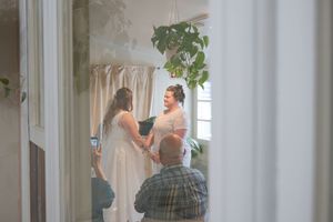 Two brides hold hands during an intimate indoor wedding ceremony with lush greenery and soft natural light.