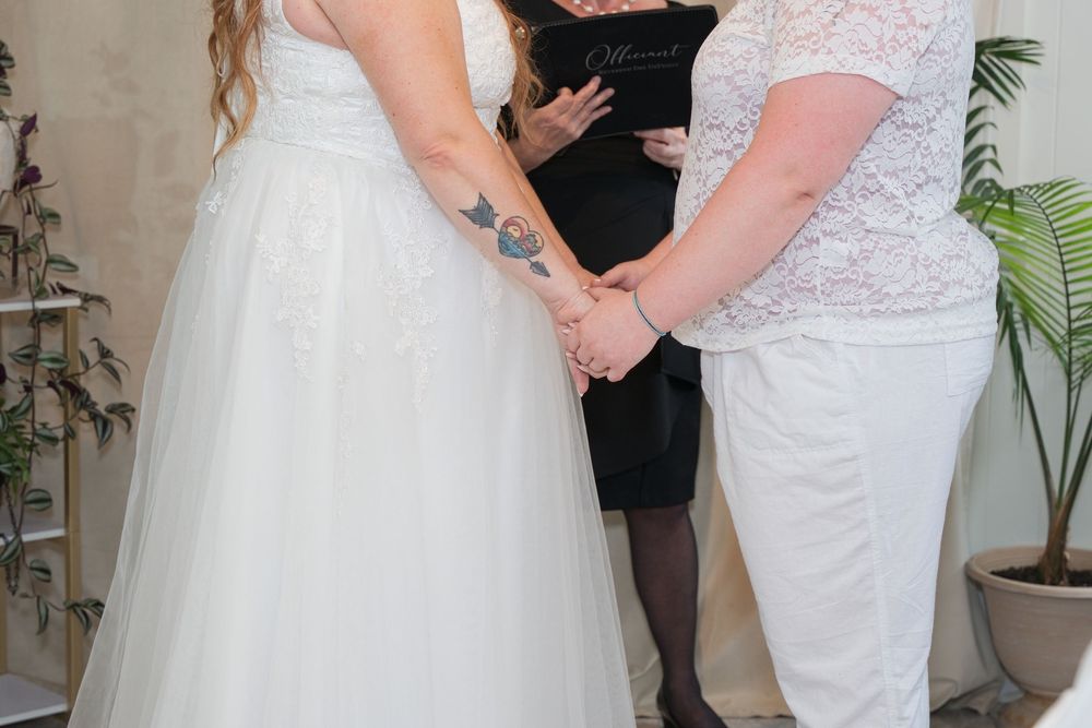 Two brides holding hands during a wedding ceremony with an officiant in the background.