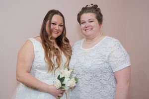 Two brides in white lace dresses smile together, one holding a bouquet of white roses at their wedding ceremony.