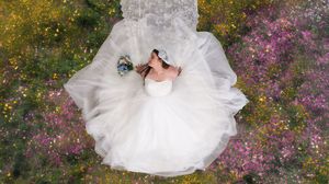 Aerial view of a bride in a white ball gown wedding dress lying among colorful wildflowers holding a bouquet.