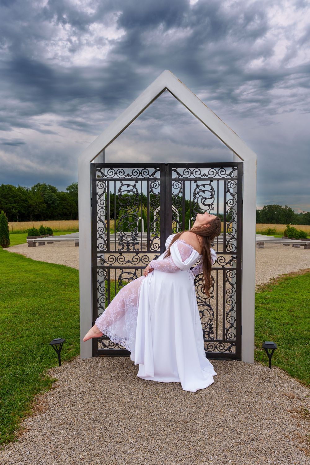 Woman in white dress posing gracefully at ornate iron garden gate under dramatic cloudy sky.