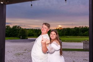 Couple in white outfits posing playfully at sunset outdoors near a rustic venue with trees in background.