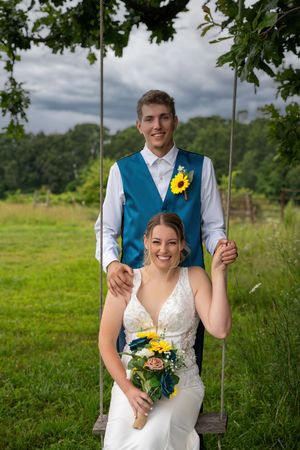 Bride sitting on rope swing holding sunflower bouquet with groom standing behind her outdoors.