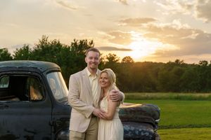 Couple embracing at sunset beside a vintage truck in a green field, golden hour light glowing behind them.
