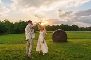 Bride and groom dancing at sunset in a green field with a hay bale, golden light glowing behind them.