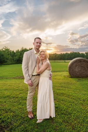 Couple posing in a green field at sunset with hay bale, man in tan suit and woman in white dress.