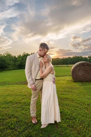 Couple embracing in a green field at sunset with hay bale, man in tan suit, woman in white dress.
