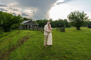 Couple embracing in lush green field at sunset with rustic barn and dramatic cloudy sky in background.