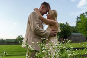A couple shares a romantic embrace outdoors, surrounded by white wildflowers, dressed in elegant wedding attire.