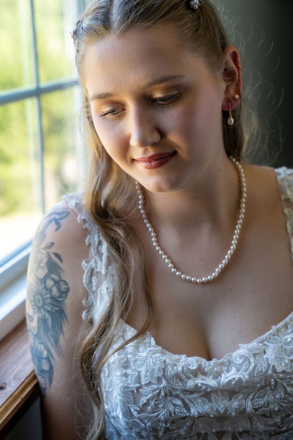 Blonde bride wearing pearl necklace and earrings in white lace dress with floral arm tattoo.