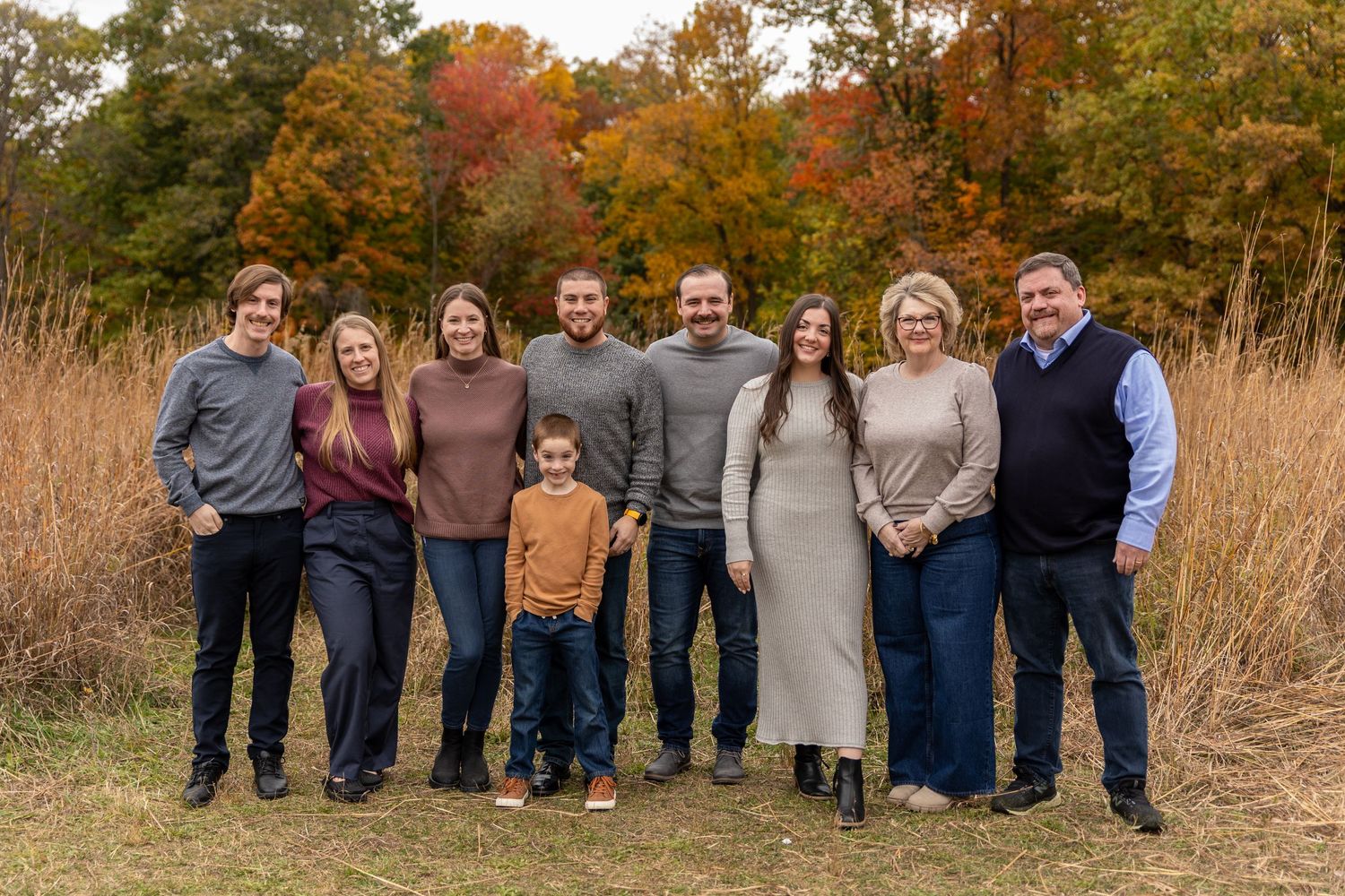 Extended family of nine posing together in an autumn field surrounded by colorful fall foliage.