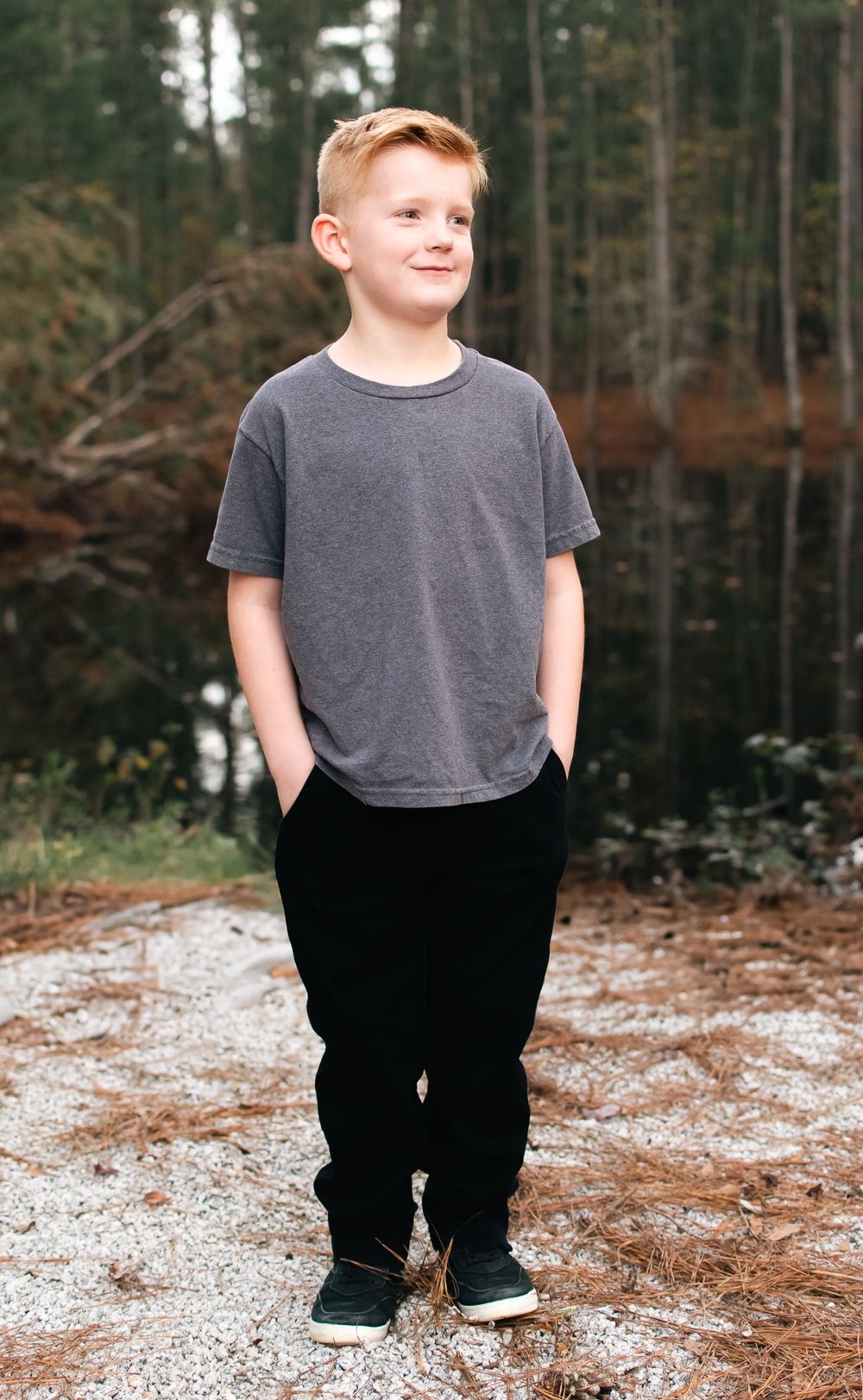 Young person in gray shirt and black pants standing on forest path.