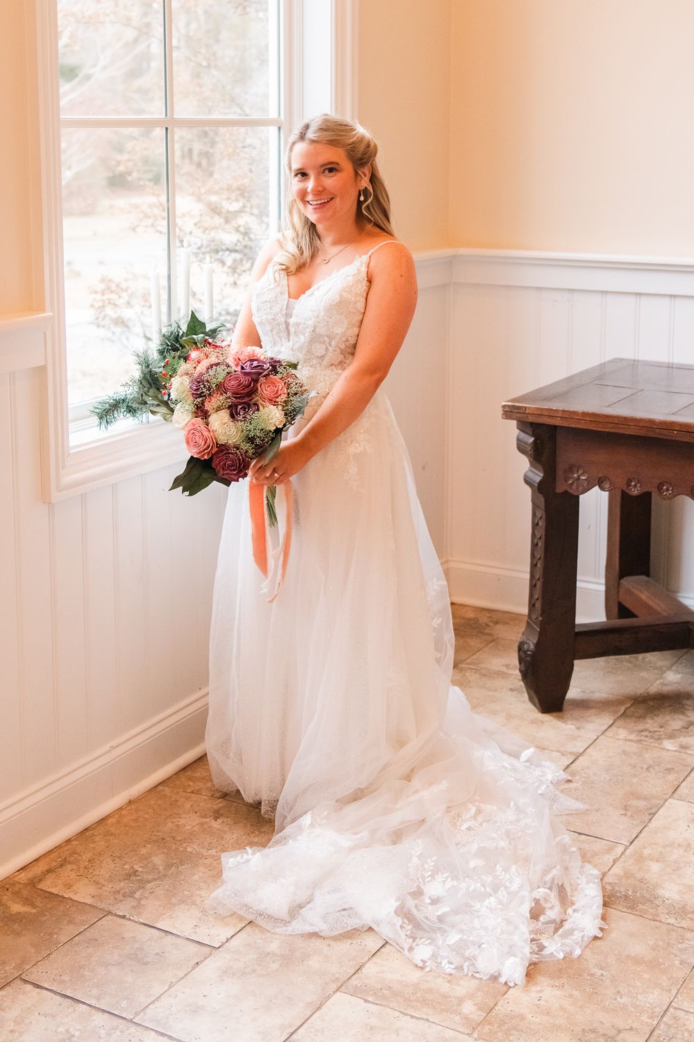 Bride in lace wedding dress holding floral bouquet in sunlit room.