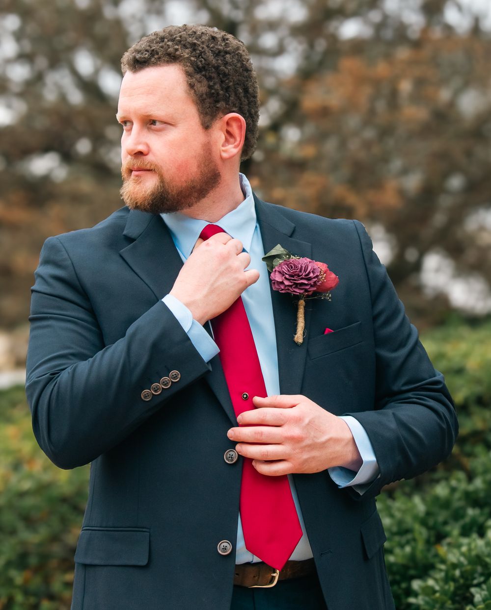 A groom in a navy suit adjusts his bright red tie while wearing a burgundy boutonniere.