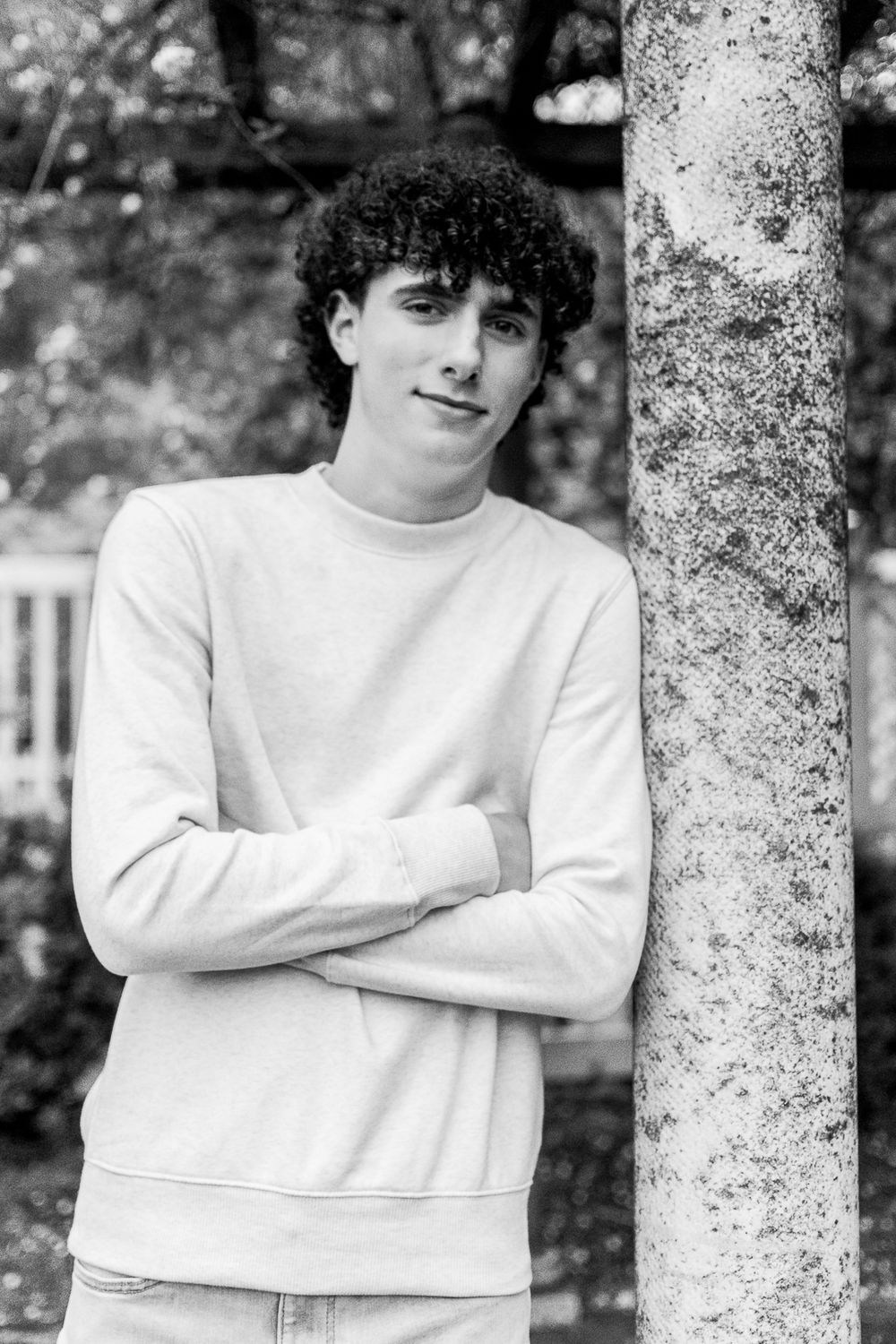 Black and white portrait against tree trunk wearing white sweater with crossed arms.