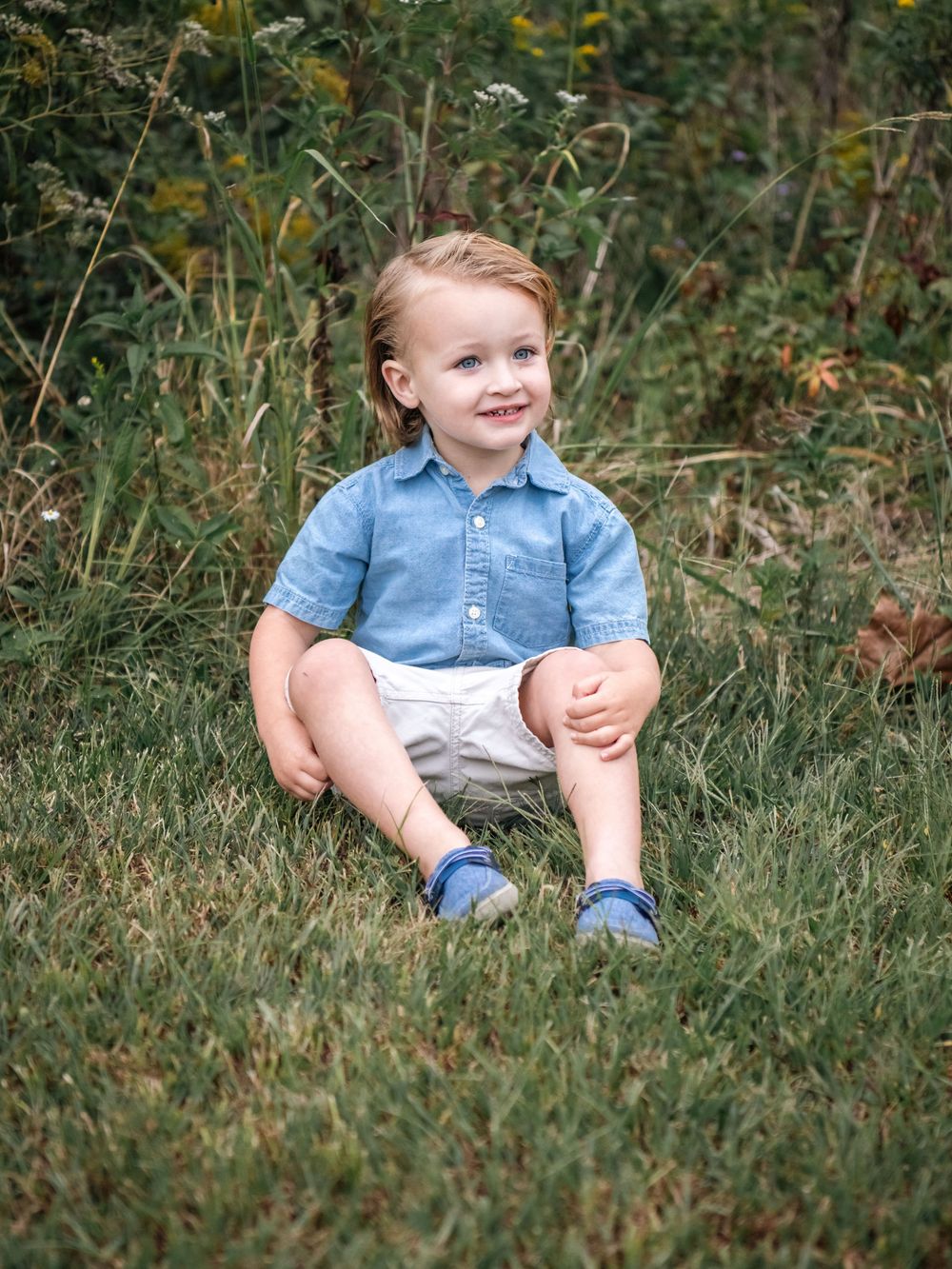 Child sitting in casual denim on grass surrounded by natural foliage.