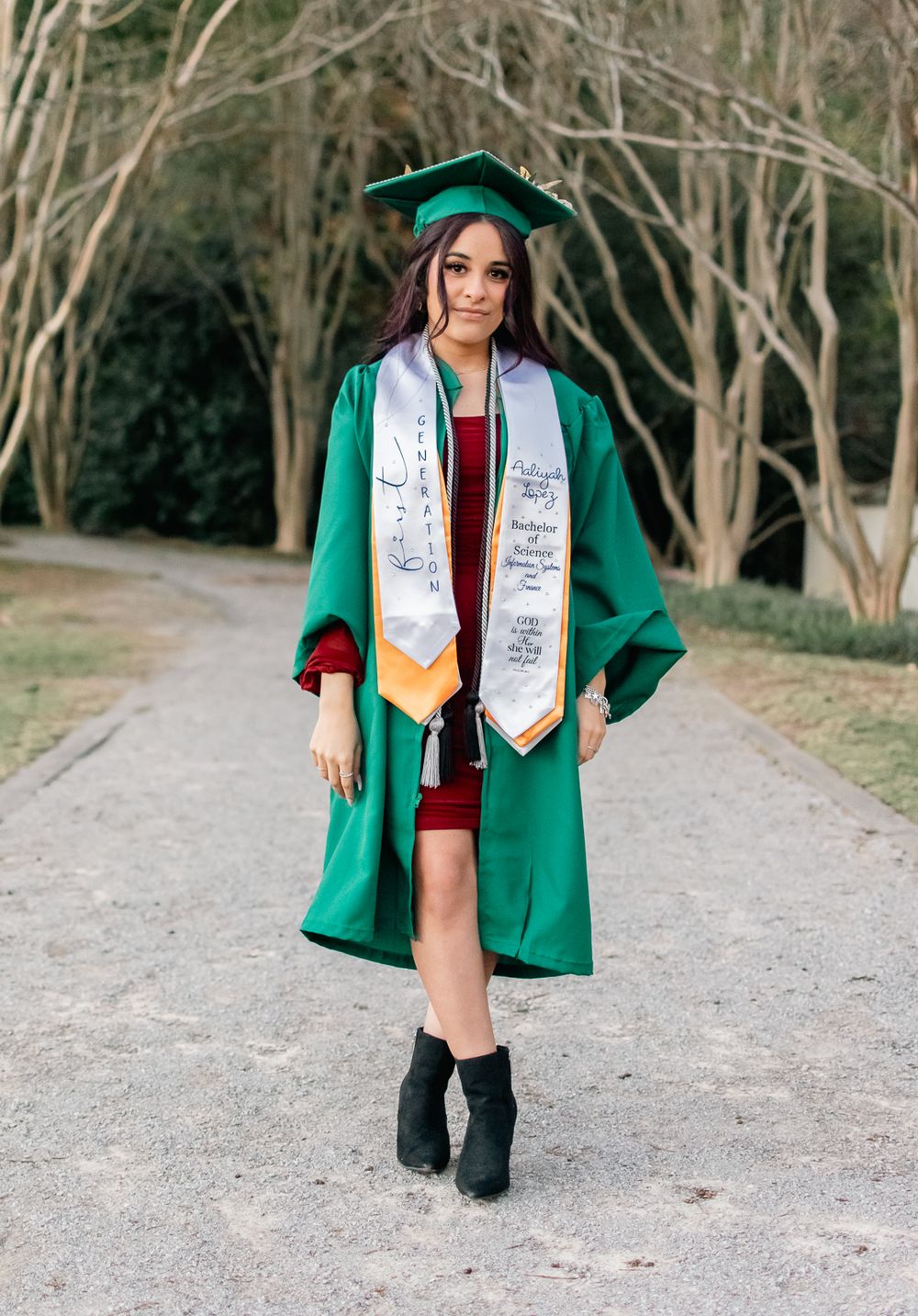 Graduate in emerald green cap and gown walking on a gravel path during photo session.